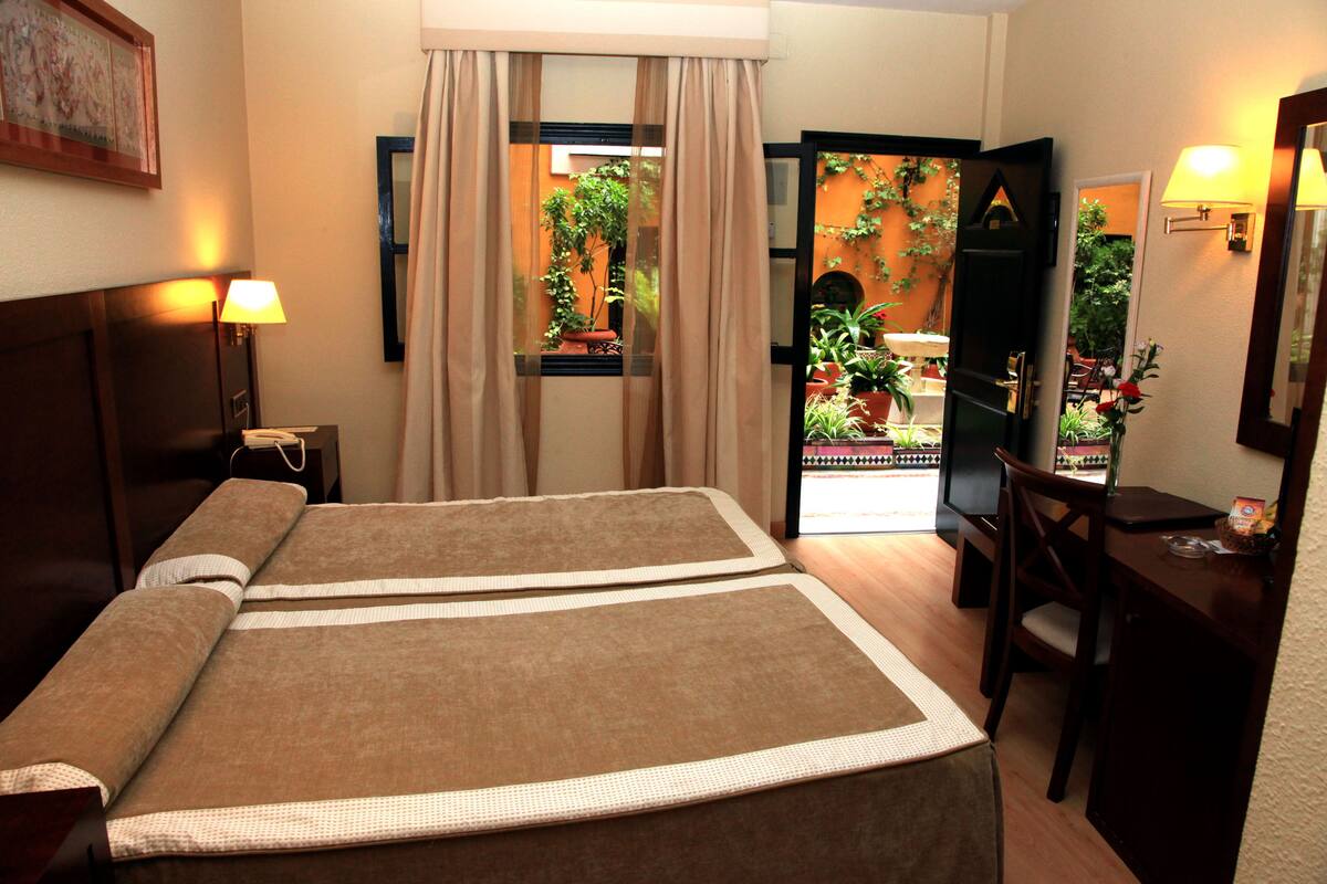 A hotel room with a bed, bedside table, lamp, and a view of a garden through a window.
