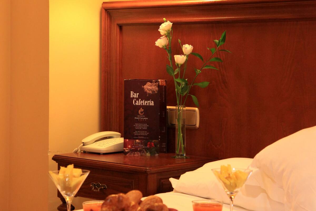 A hotel room with a neatly made bed, a bedside table with a lamp, a menu for the bar and cafeteria, and a vase with flowers.