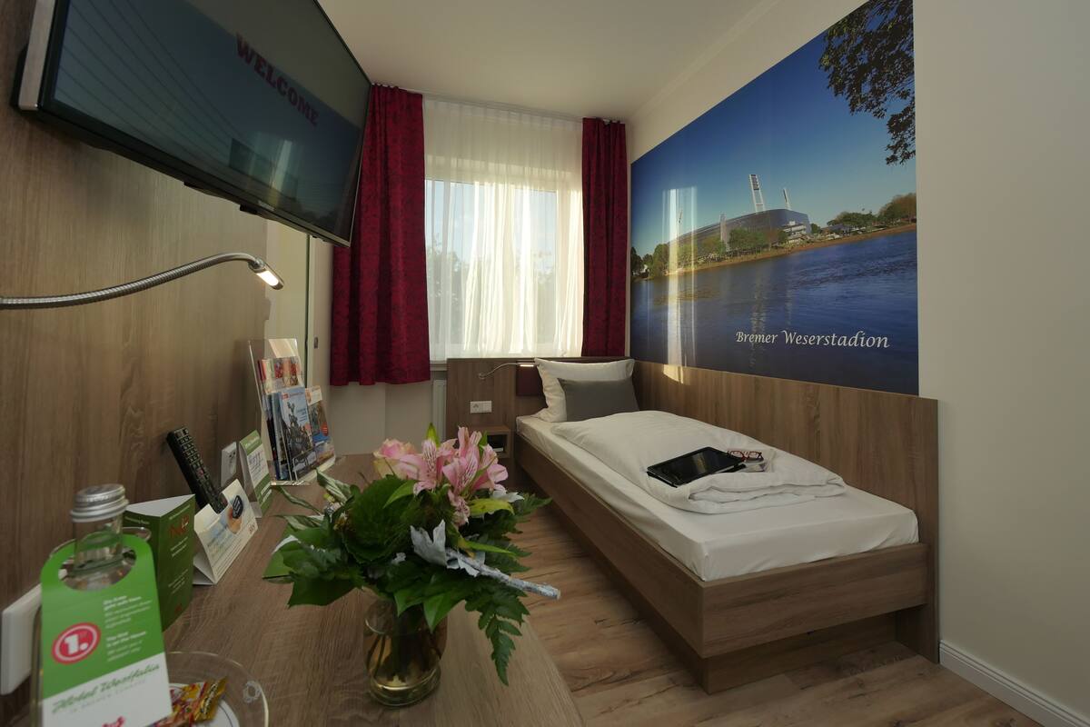 A modern hotel room with a bed, a desk with a lamp, a vase of flowers, a poster of a bridge, and a TV on the wall.