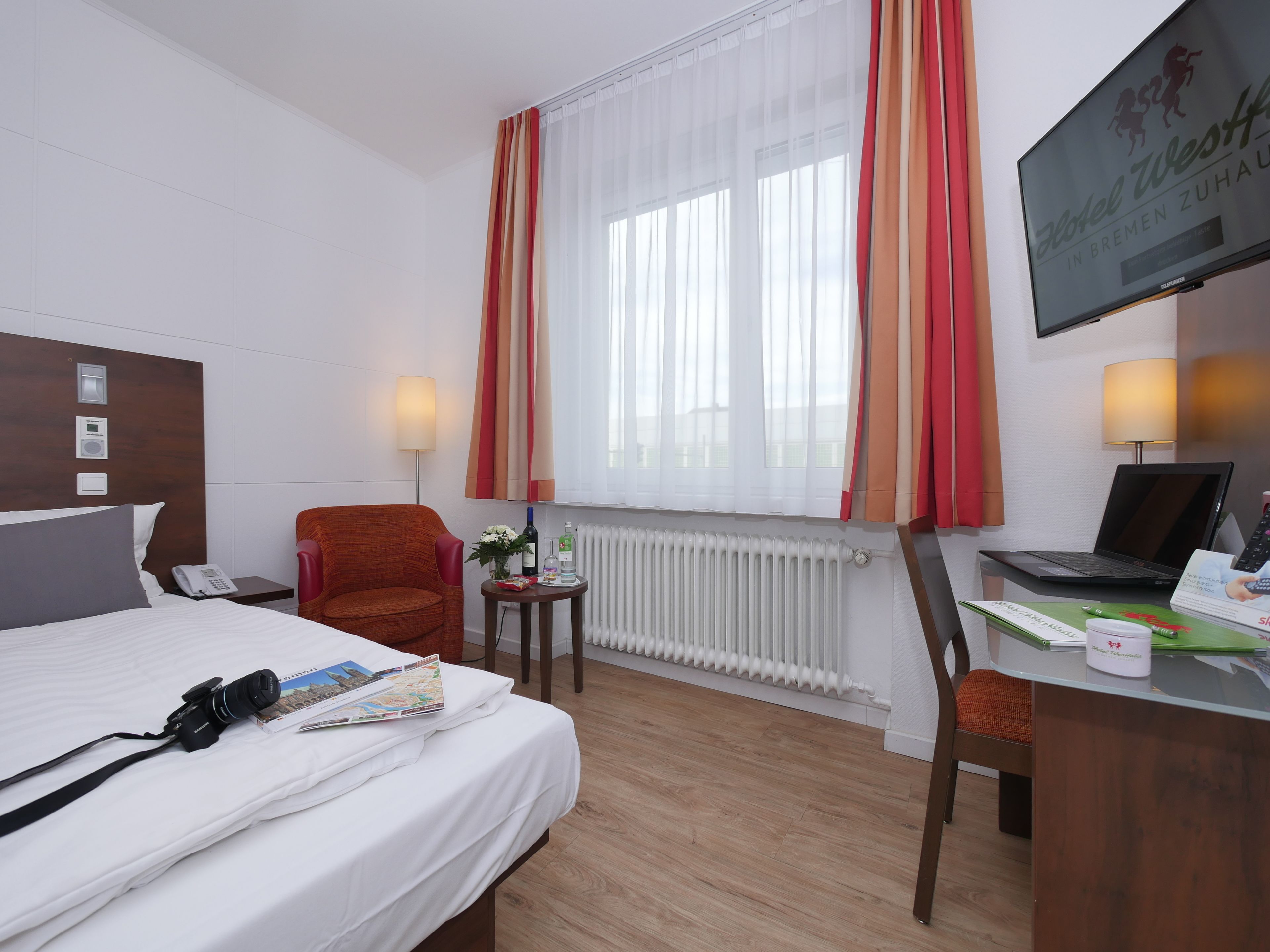 Ett hotellrum med en säng, ett skrivbord med en bärbar dator, en stol, ett element, ett fönster med gardiner och en tv på väggen.
