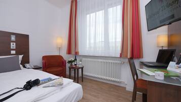 Ett hotellrum med en säng, ett skrivbord med en bärbar dator, en stol, ett element, ett fönster med gardiner och en tv på väggen.