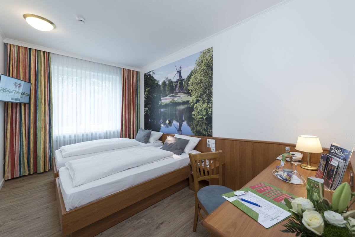 A hotel room with a bed, a desk with a lamp, a chair, a window with curtains, and a wall with a scenic painting.