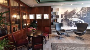 Ein gemütliches Zimmer mit einer holzverkleideten Wand, einem runden Tisch mit Stühlen, einem Bücherregal und einem großen Fenster mit Blick auf eine Straßenszene.
