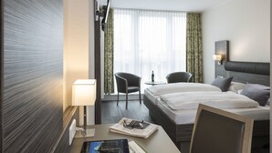 Ein Hotelzimmer mit einem Bett, zwei Sesseln, einem Schreibtisch mit Lampe und einem fensterbepflanzten Fenster.