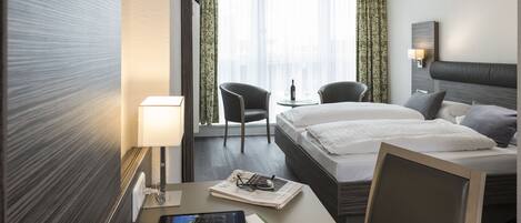 A hotel room with a bed, two chairs, a desk with a lamp, and a window with curtains.