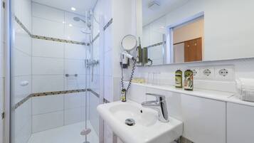 Un baño moderno con ducha envuelta en vidrio, un lavabo blanco con espejo y un estante con artículos de aseo.