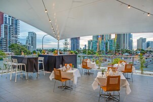 An outdoor dining area with tables and chairs, a city skyline in the background, and a view of a body of water.