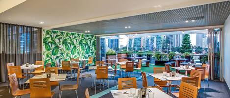 Een restaurant met meerdere tafels en stoelen, een groene wand met bladeren en uitzicht op hoge gebouwen.