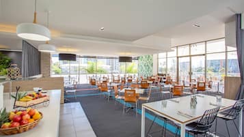 A modern dining area with multiple tables and chairs, large windows offering city views, and a fruit basket on the counter.