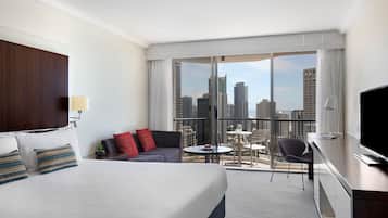 Une chambre d’hôtel avec un grand lit, un canapé, une petite table, une chaise et un téléviseur. La chambre dispose d’un balcon avec vue sur la ville.