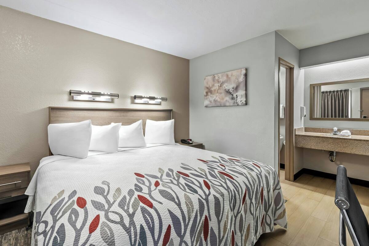 Habitación de hotel con cama, mesita de noche, tocador y baño con espejo, lavabo y toallero.