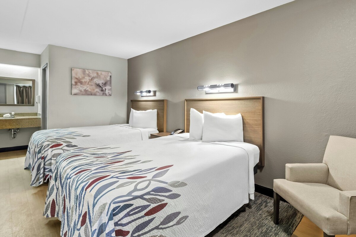 A hotel room with two beds, a chair, a mirror, and a wall-mounted light fixture.