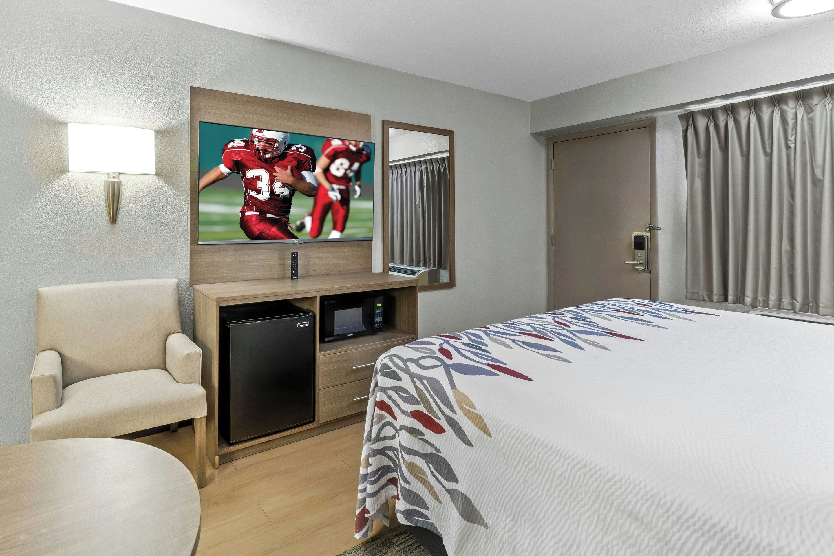 A hotel room with a bed, a TV mounted on the wall, a desk with a refrigerator, a chair, and a window with curtains.