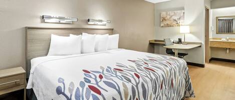 Habitación de hotel con cama, mesita de noche, lámpara y baño con lavamanos y espejo.