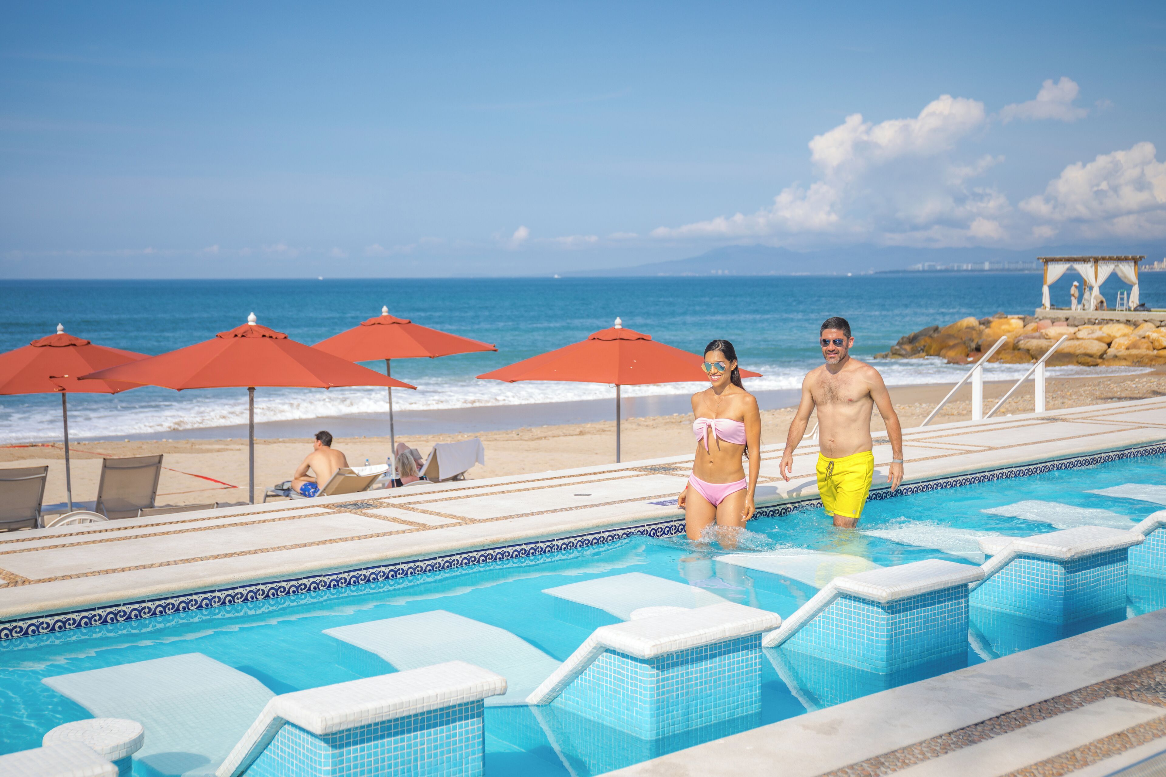 A pool area with a view of the ocean, red umbrellas, and lounge chairs.