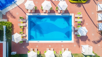 Vista aérea de una piscina rodeada de sillas de descanso y sombrillas, con suelo de ladrillo y vegetación.