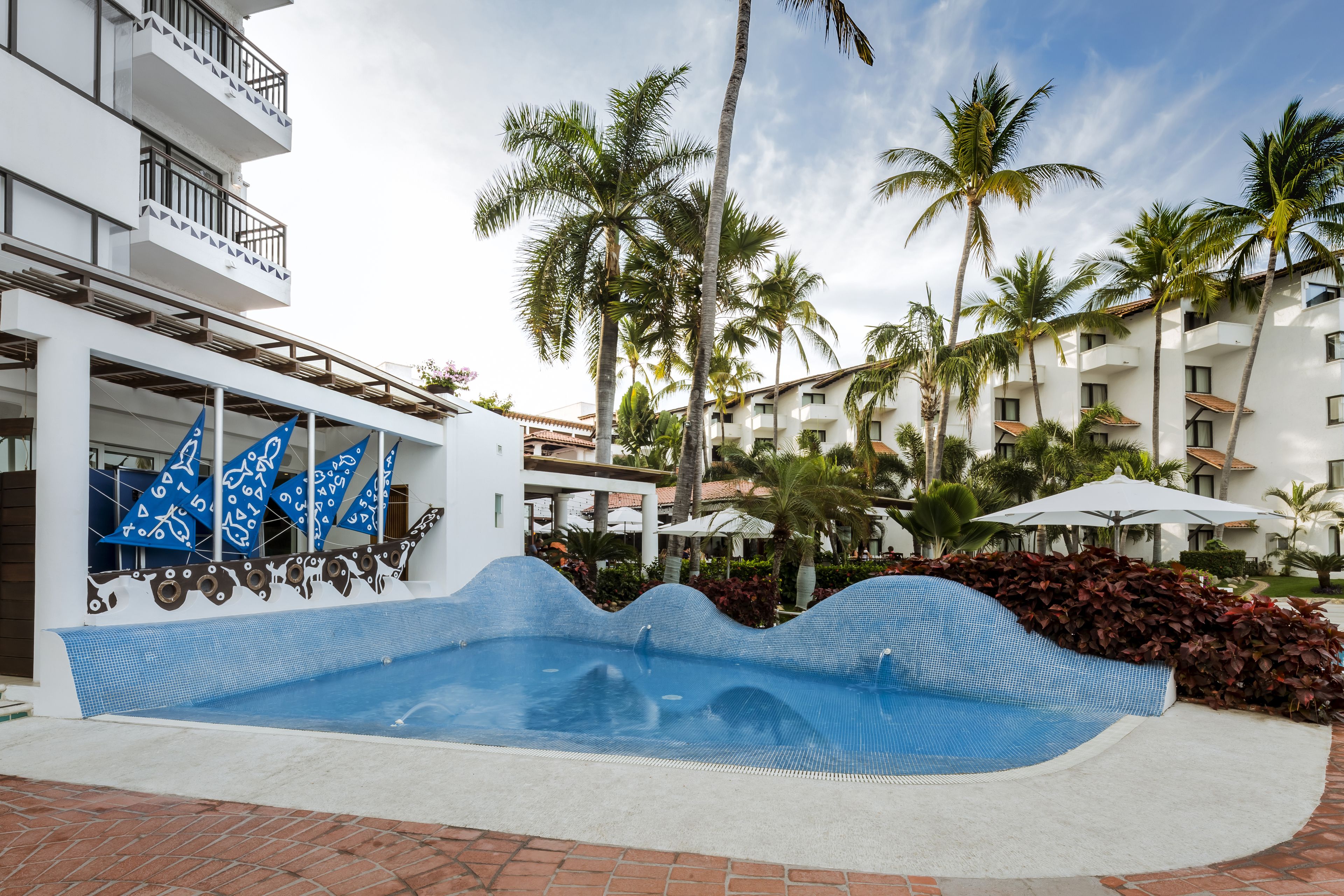 Une piscine à bords ondulés, entourée de palmiers et bordée d’un bâtiment blanc doté de balcons.