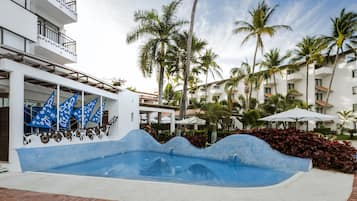A swimming pool with a wavy edge design, surrounded by palm trees and a white building with balconies.