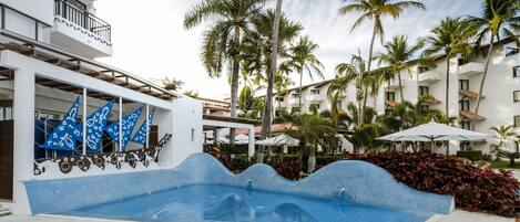 Une piscine à bords ondulés, entourée de palmiers et bordée d’un bâtiment blanc doté de balcons.