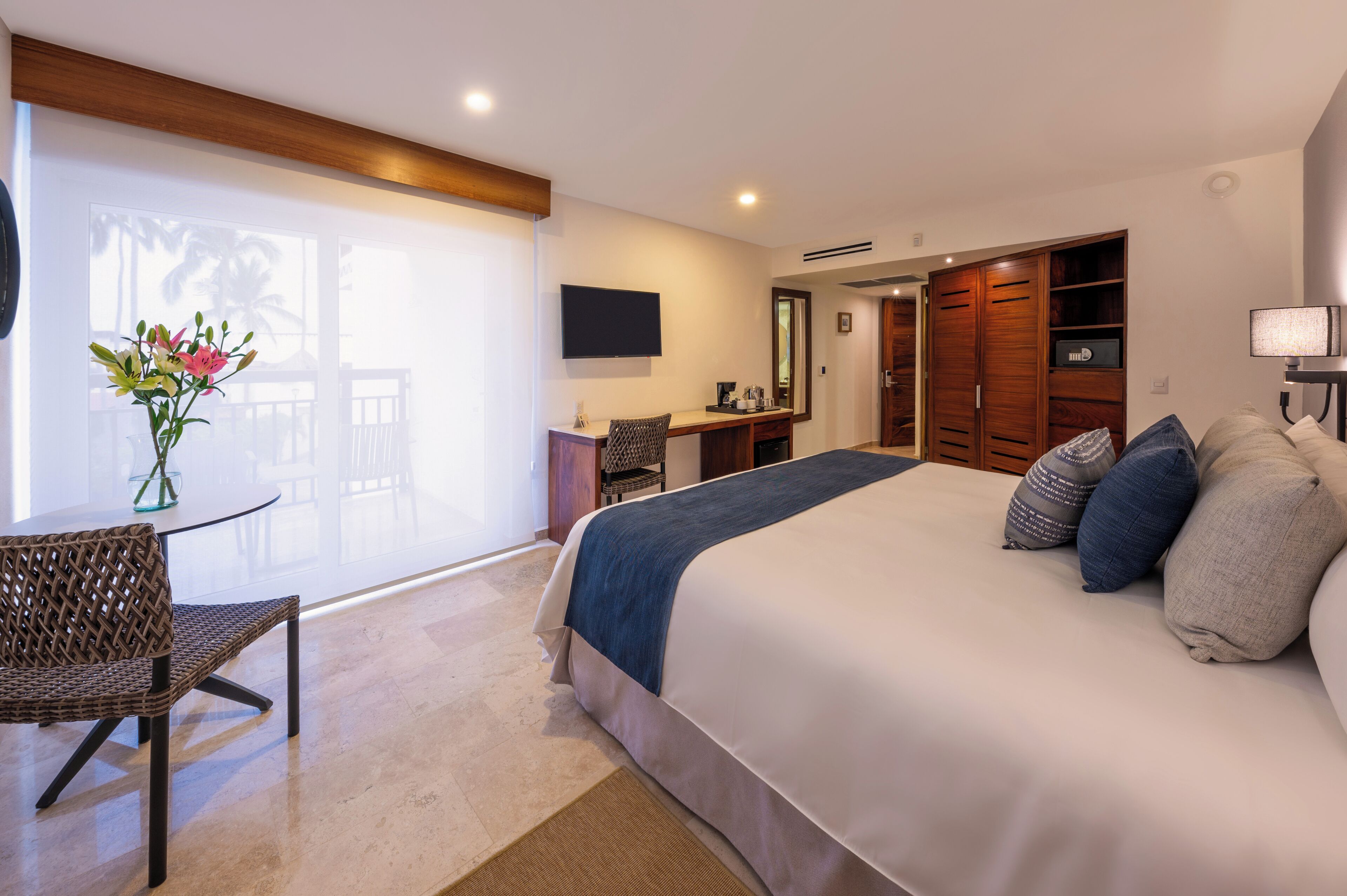 A hotel room with a large bed, a desk, a chair, a TV, and a view of palm trees.