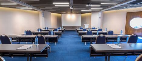 Un salón de conferencias con filas de mesas y sillas, una pantalla de proyector y un atril.