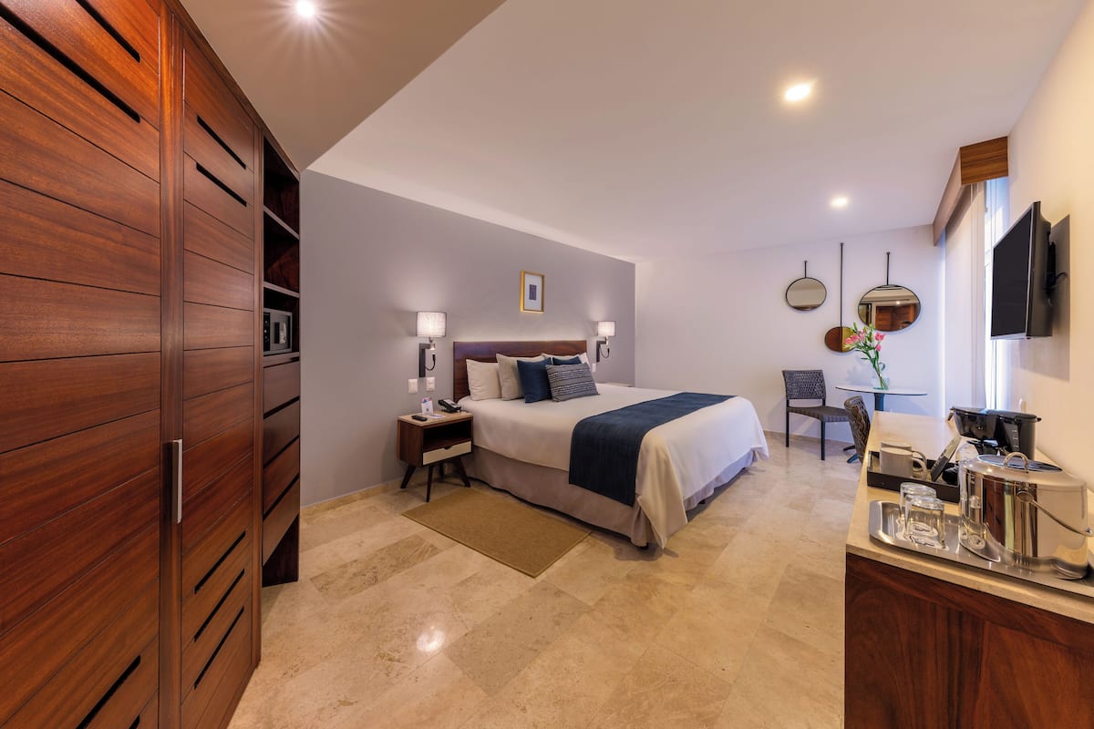 Une chambre d’hôtel moderne avec une armoire en bois, un lit recouvert d’une couette bleue et blanche, une table de chevet avec des lampes, une petite table avec une plante et un téléviseur fixé au mur.