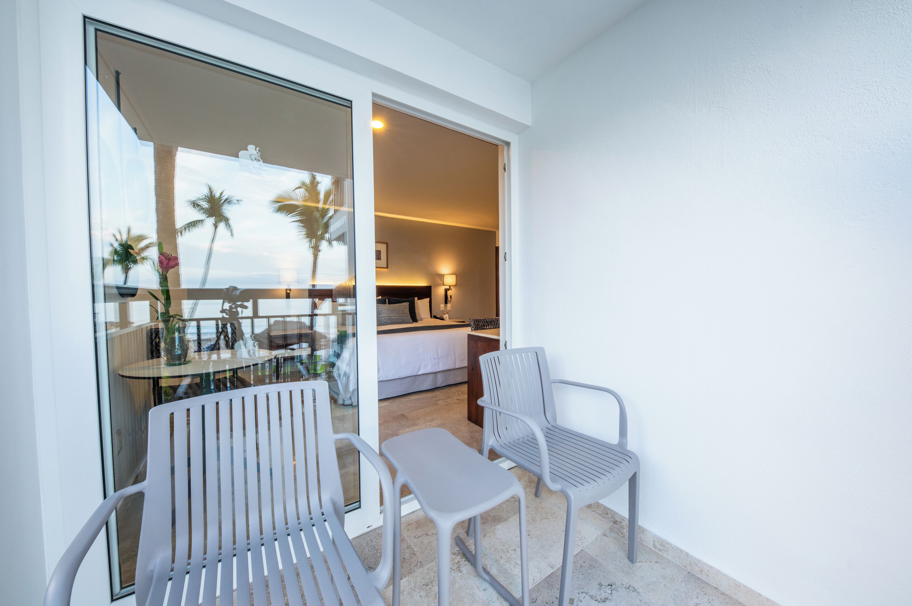 Ein Balkon mit zwei Stühlen und einem Tisch, ein Schlafzimmer mit Bett und Nachttisch sowie der Blick auf Palmen.