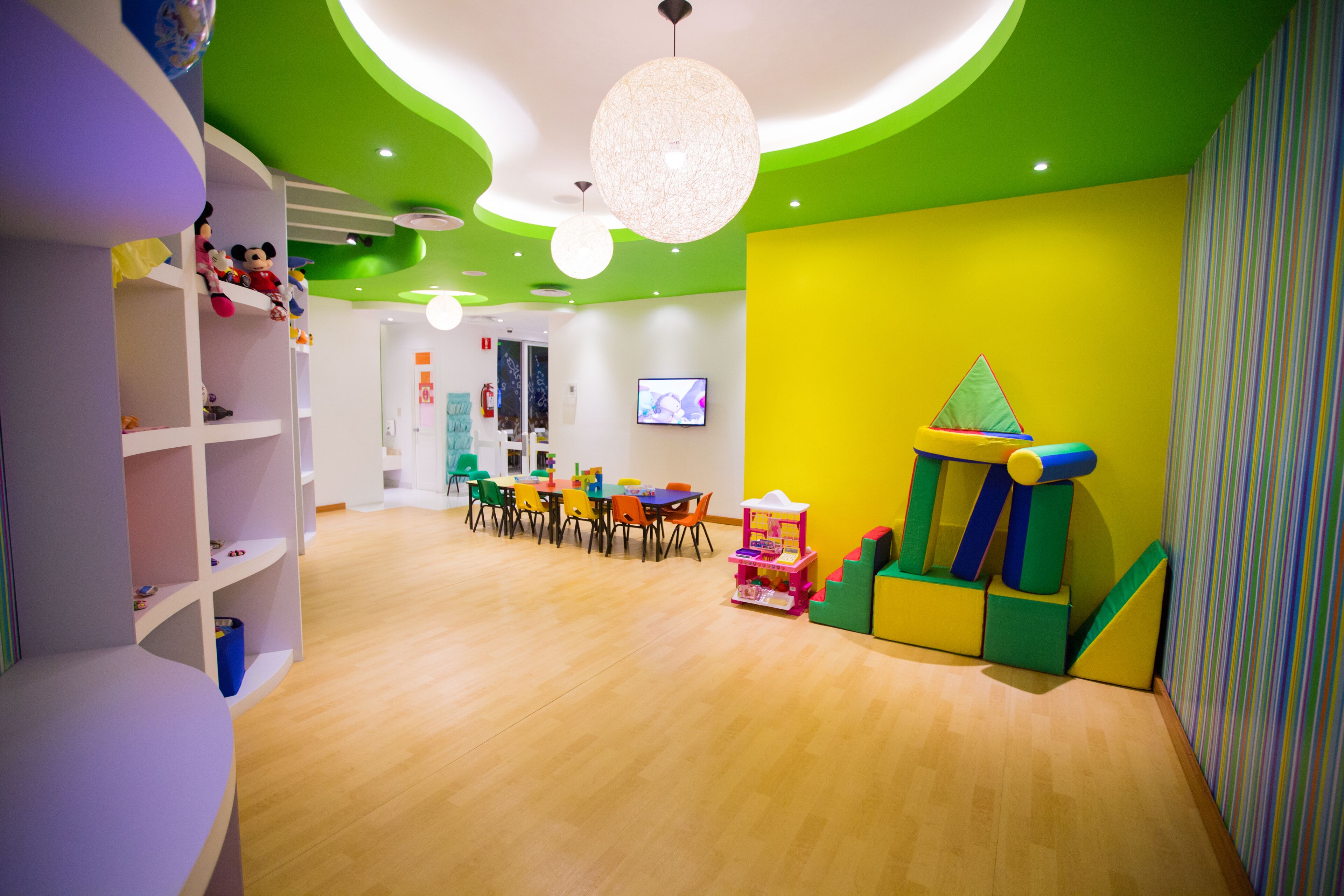 Uma sala de jogos infantil bem iluminada, com piso de madeira, estruturas de recreação coloridas e uma televisão na parede.