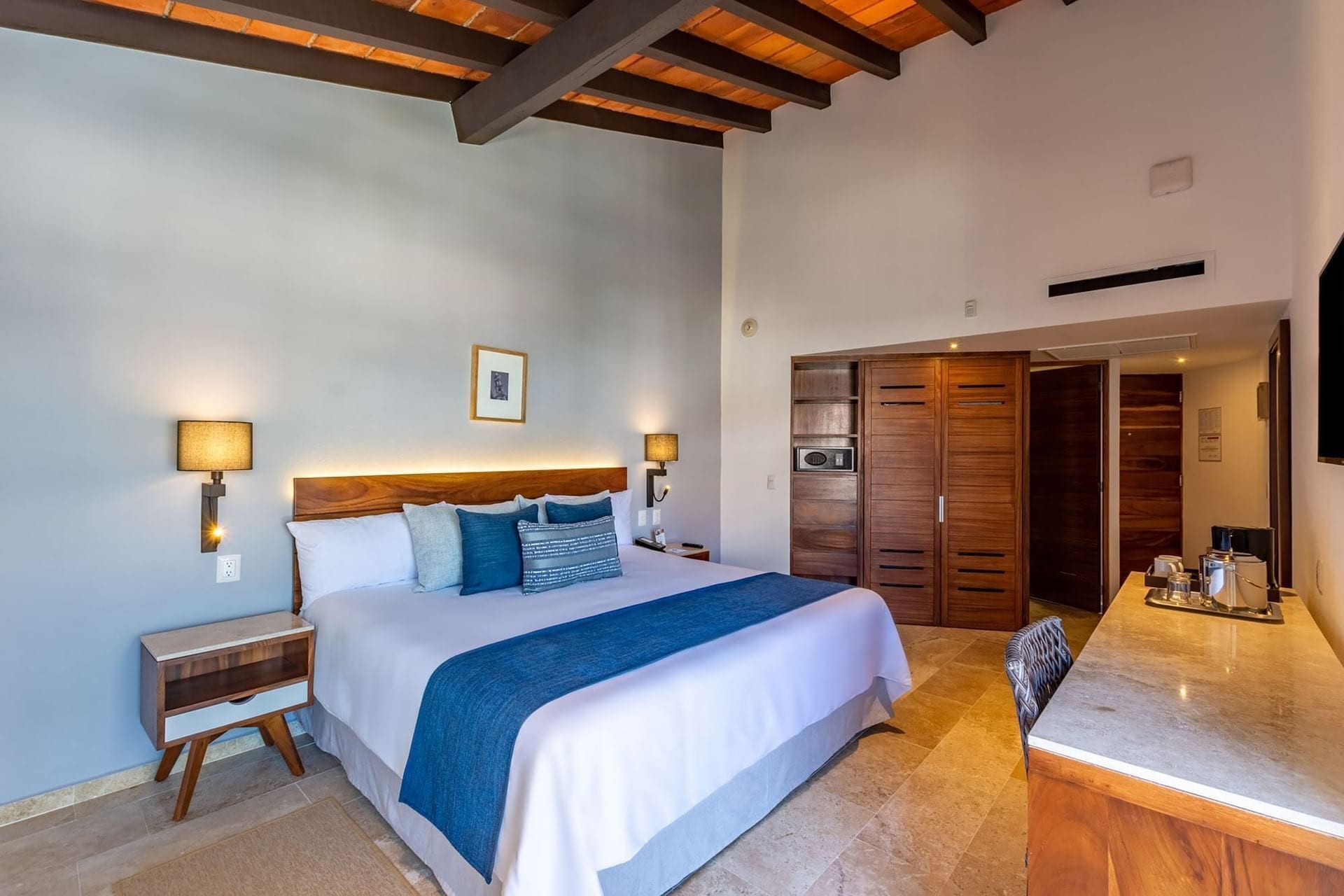 A hotel room with a bed, bedside lamps, a wooden headboard, a wardrobe, and a kitchenette.