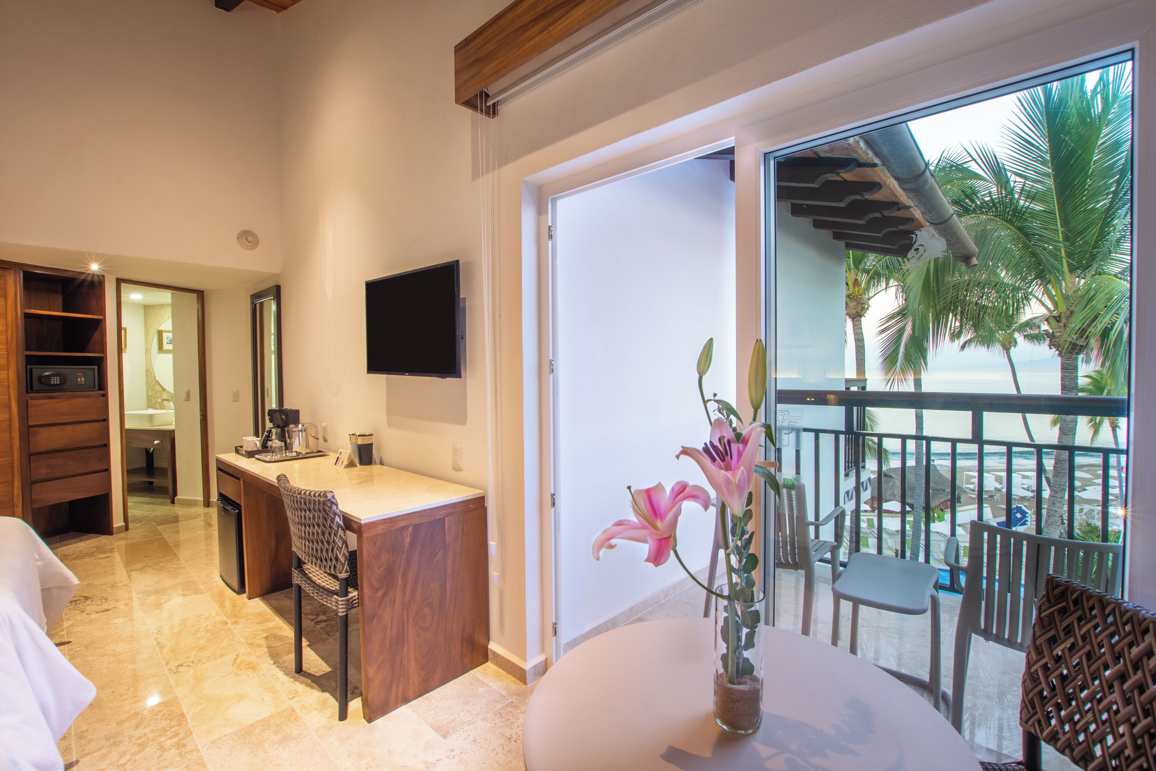 A modern hotel room with a kitchen area, a dining table with chairs, a large window with a view of palm trees and the ocean, and a flat-screen TV mounted on the wall.