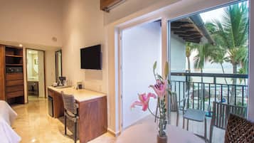 A modern hotel room with a kitchen area, a dining table with chairs, a large window with a view of palm trees and the ocean, and a flat-screen TV mounted on the wall.