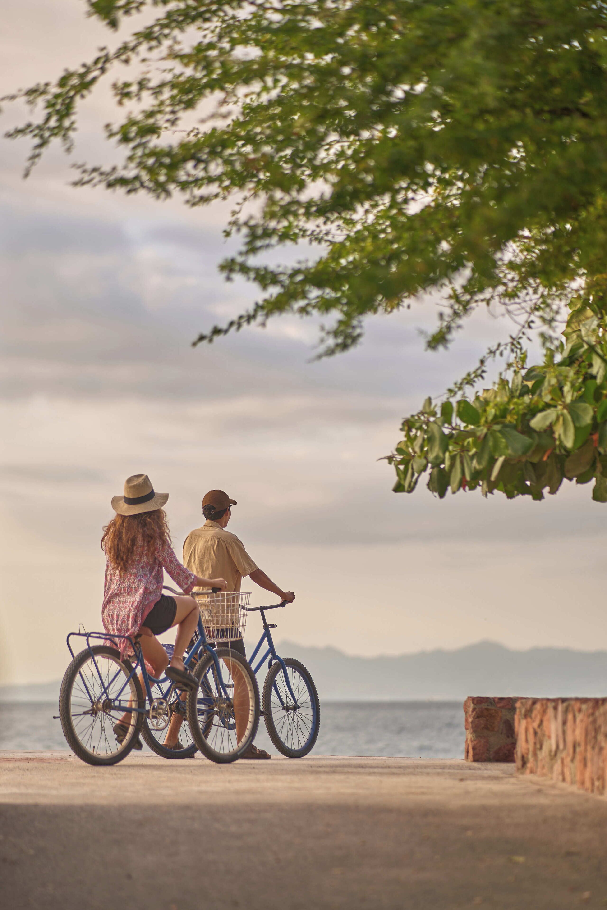 Two people on a tandem bicycle with a basket, overlooking a body of water and distant mountains.