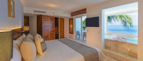 A bedroom with a large bed, a TV, a balcony with a view of the ocean, and a bathroom with a bathtub.