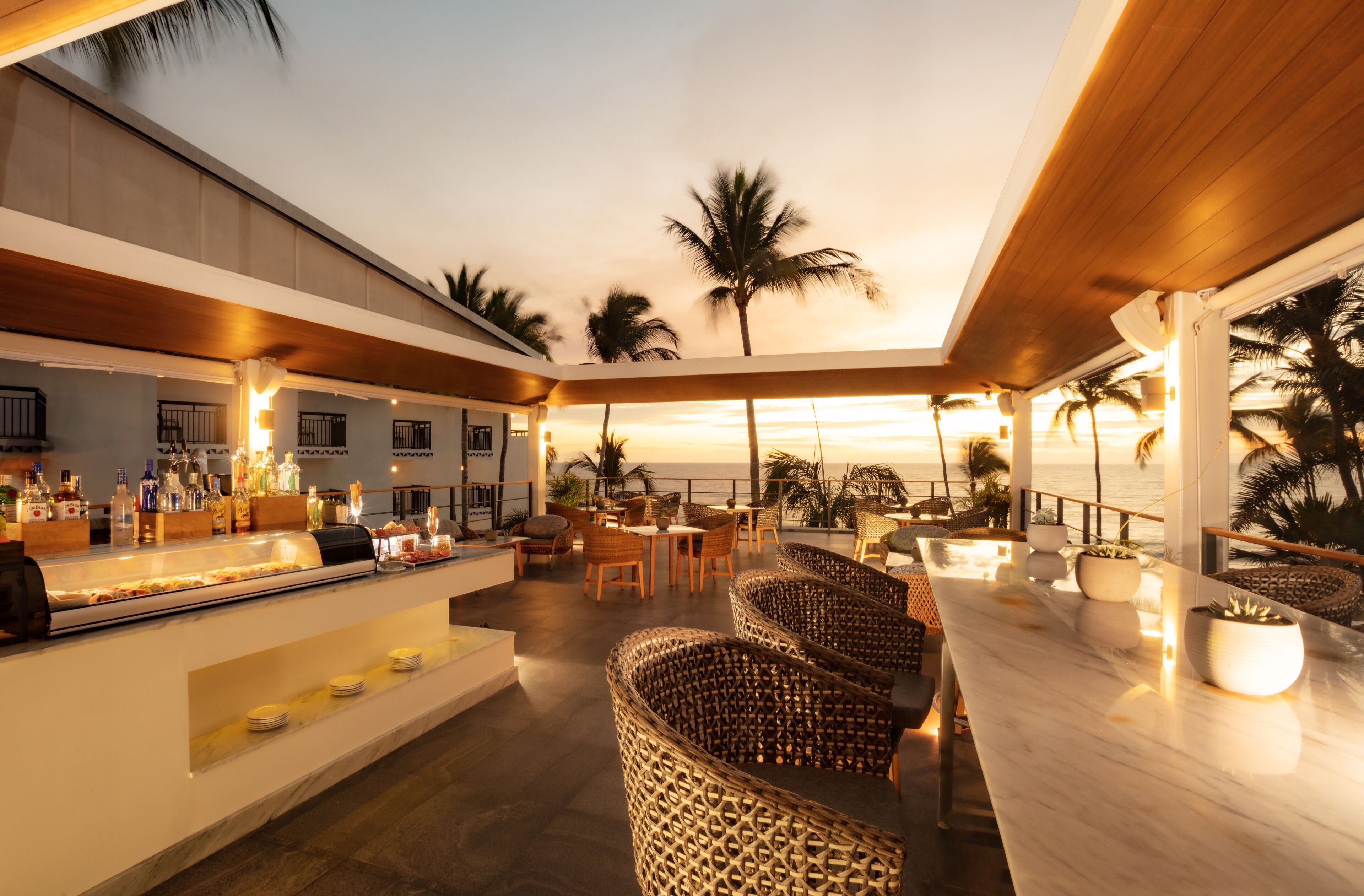 A rooftop bar with a long counter, wicker chairs, and a view of the ocean at sunset.