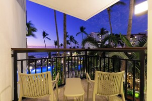 Un balcón con dos sillas y una mesa, con vista a una piscina y palmeras bajo un cielo crepuscular.