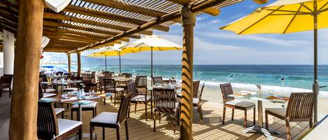 A beachside restaurant with outdoor seating, wooden tables, and yellow umbrellas.
