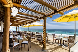 Un restaurante frente a la playa con terraza, mesas de madera y sombrillas amarillas.