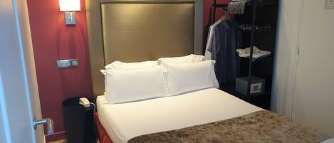 Una habitación de hotel con cama, mesita de noche, lámpara, armario y cesto de basura.