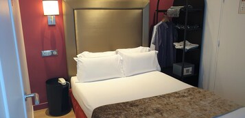 A hotel room with a bed, a bedside table, a lamp, a wardrobe, and a trash bin.