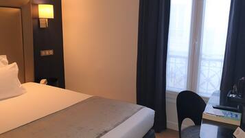 A hotel room with a bed, a desk with a chair, a lamp, and a window with curtains.