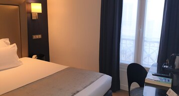 Una camera d'albergo con un letto, una scrivania con una sedia, una lampada e una finestra con tende.