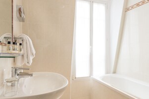 A bathroom with a large white sink, a glass shelf with toiletries, a towel rack, and a bathtub.