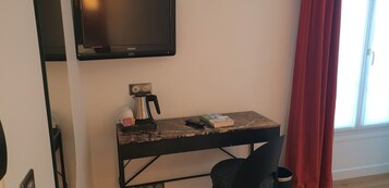 A small room with a TV mounted on the wall, a desk with a coffee maker, a book, and a remote control.