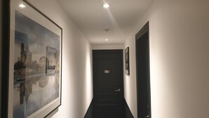 Un pasillo con un cuadro de un paisaje urbano enmarcado, una puerta con un número y una imagen enmarcada en la pared.