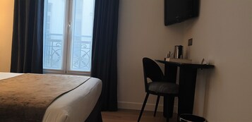 A hotel room with a bed, a desk with a chair, a television mounted on the wall, and a view of a balcony through the window.