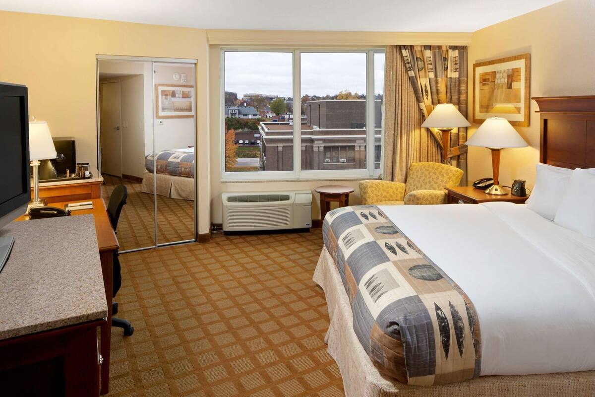 Ein Hotelzimmer mit Bett, Schreibtisch, Stuhl und Blick auf die Stadt durch ein großes Fenster.