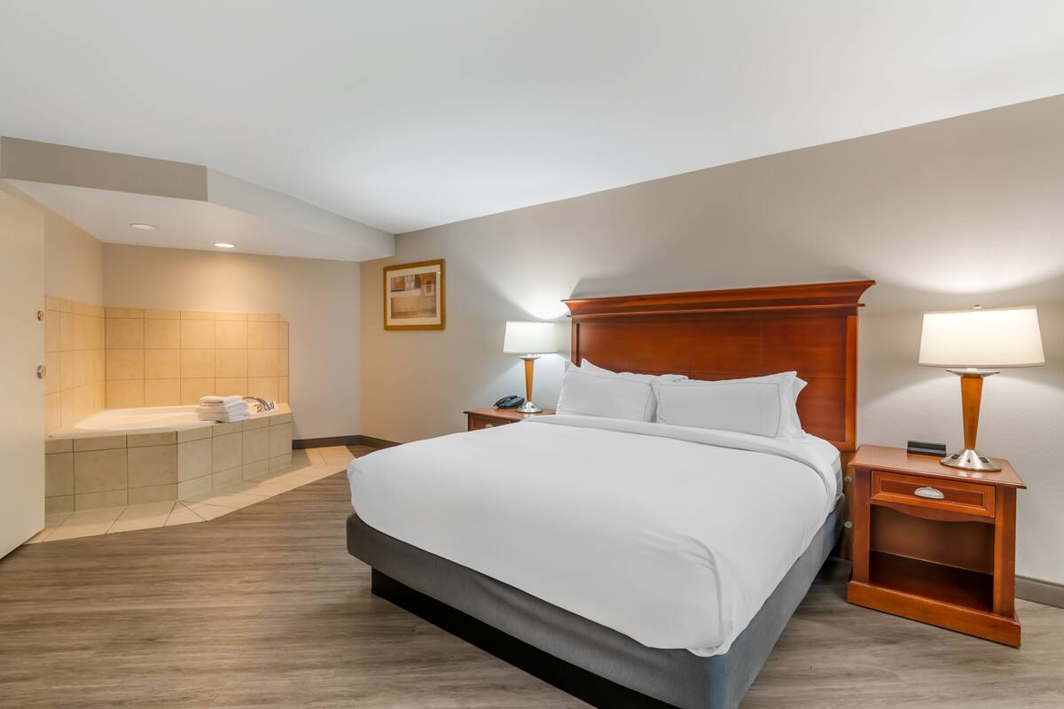 Ein Hotelzimmer mit einem großen Bett, einem hölzernen Kopfteil, einem Nachttisch mit Lampe, einem Badezimmer mit Badewanne und einem gerahmten Bild an der Wand.