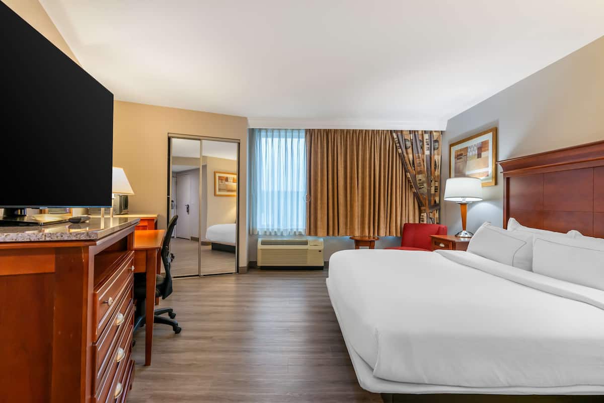 Ein Hotelzimmer mit einem großen Bett, einem Schreibtisch mit Flachbildfernseher, einem Sessel, einer Schreibtischlampe, einem Fenster mit Vorhängen und einem Kleiderständer.
