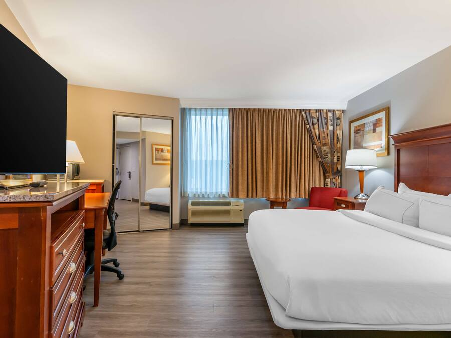 A hotel room with a large bed, a desk with a flat-screen TV, a chair, a desk lamp, a window with curtains, and a coat rack.