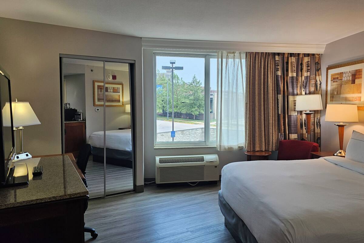 Ein Hotelzimmer mit Bett, Schreibtisch mit Lampe, Stuhl, Spiegel, Fenster mit Vorhängen und Blick nach außen.